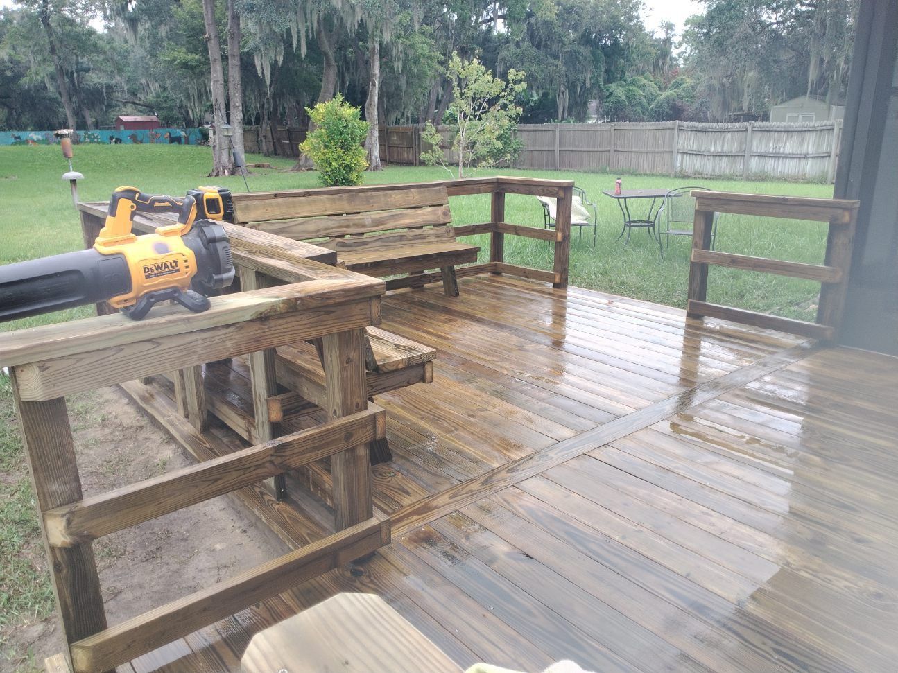 Person using a leaf blower on a wet wooden deck in a backyard.