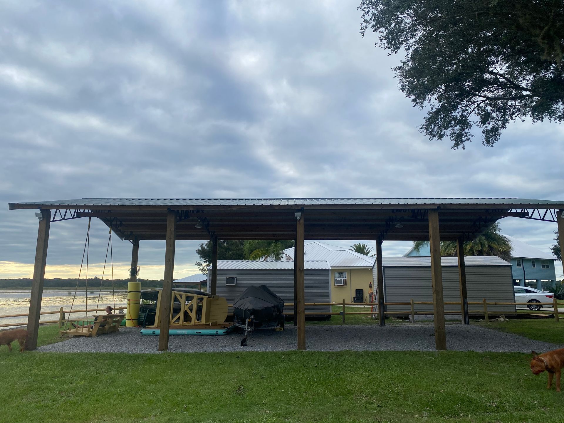 Covered outdoor area with gravel, wooden posts, and a corrugated metal roof, overlooking water.