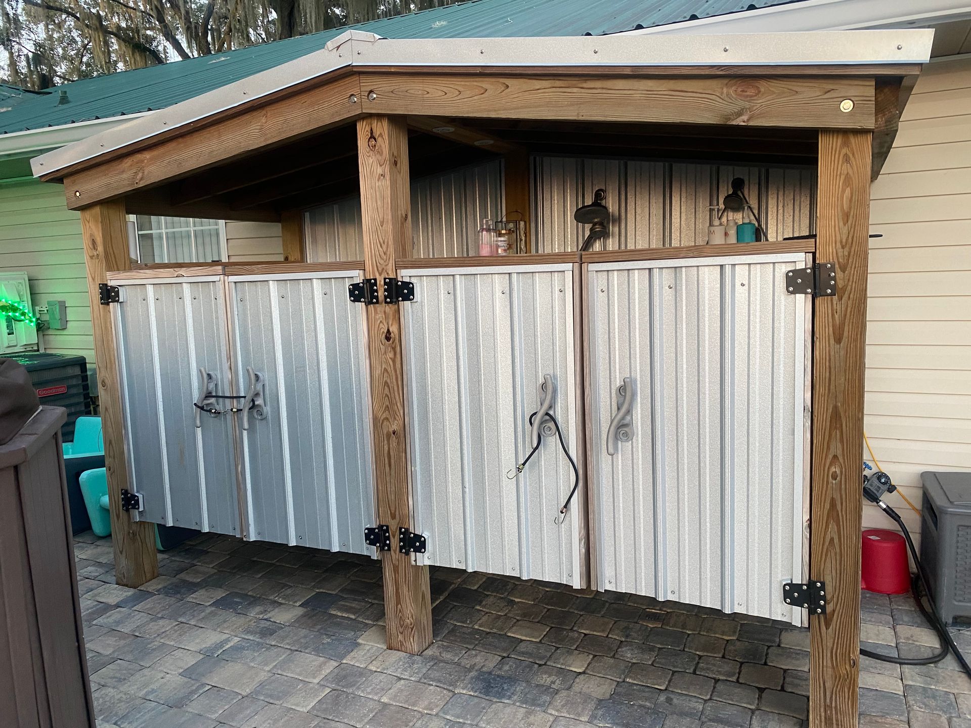 Outdoor shower with corrugated metal doors, wooden frame and roof, on a paved patio.