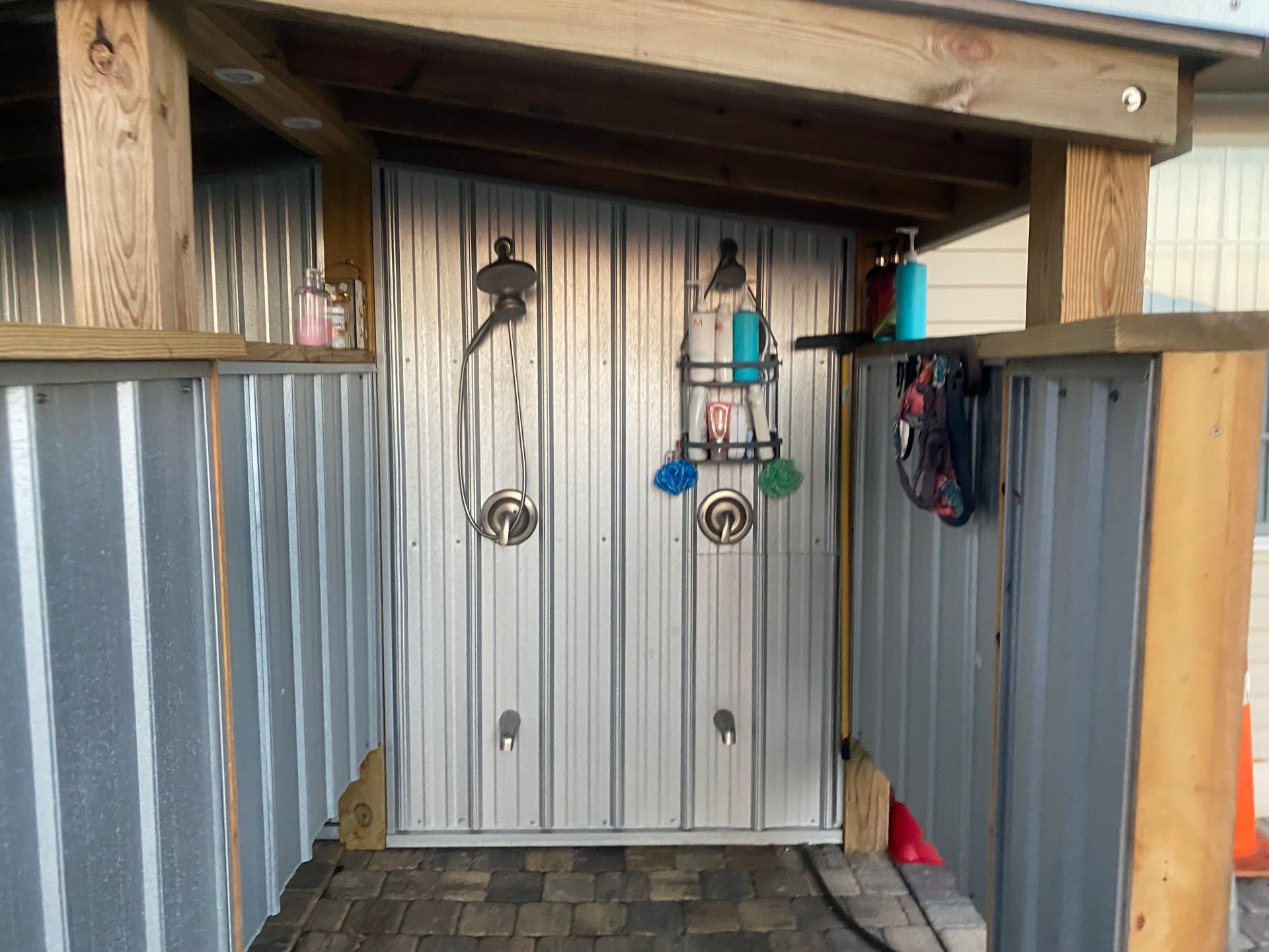 Outdoor shower with corrugated metal walls, two shower heads, and shelves with toiletries.