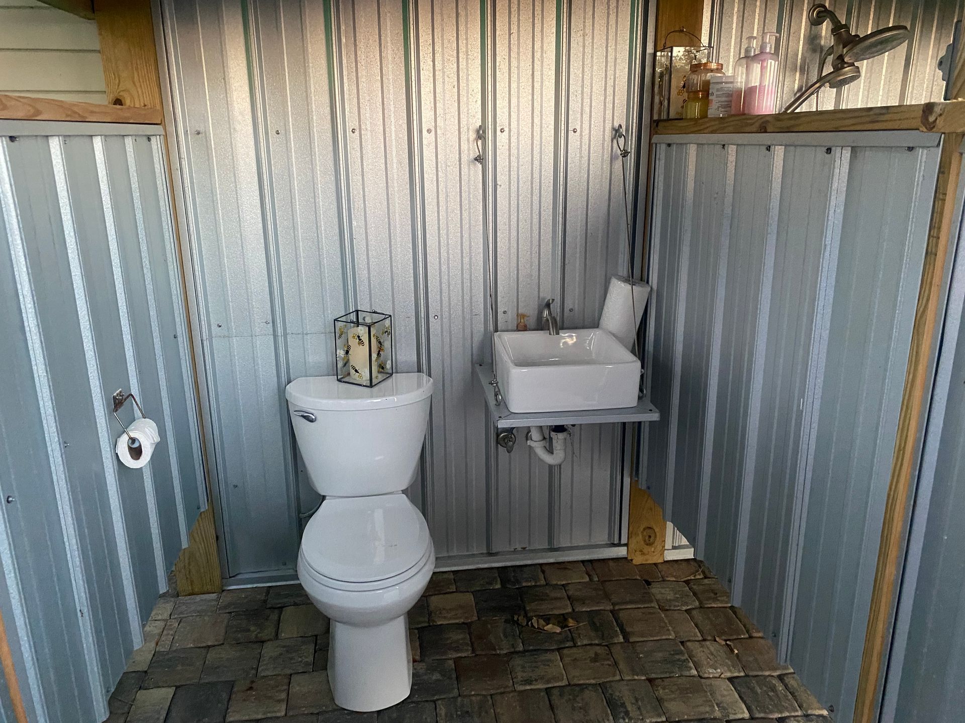 Outdoor bathroom with a toilet, sink, and corrugated metal walls. Cobblestone floor.