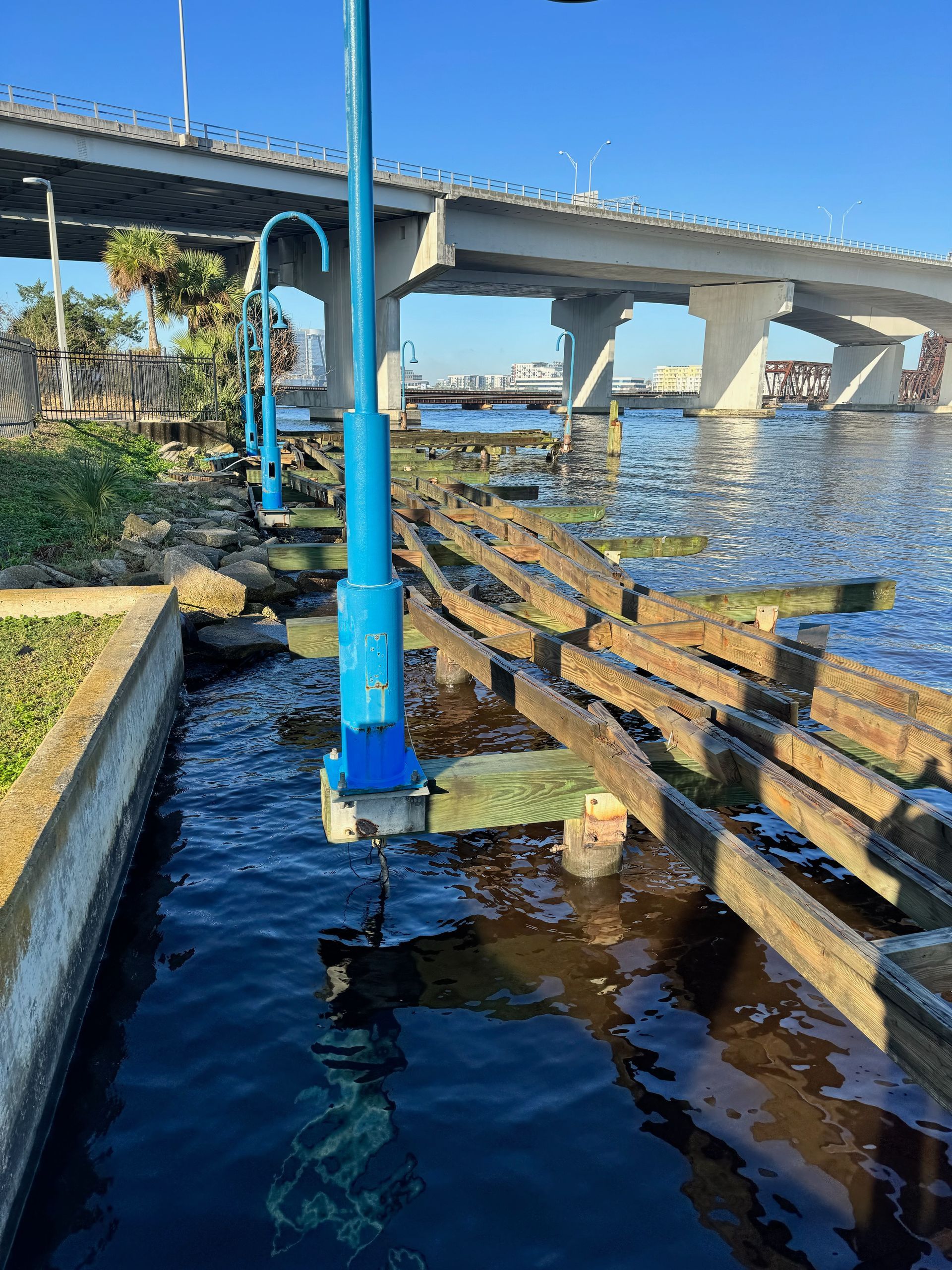 Waterfront scene with blue light poles, a dilapidated wooden dock, and a bridge overhead.