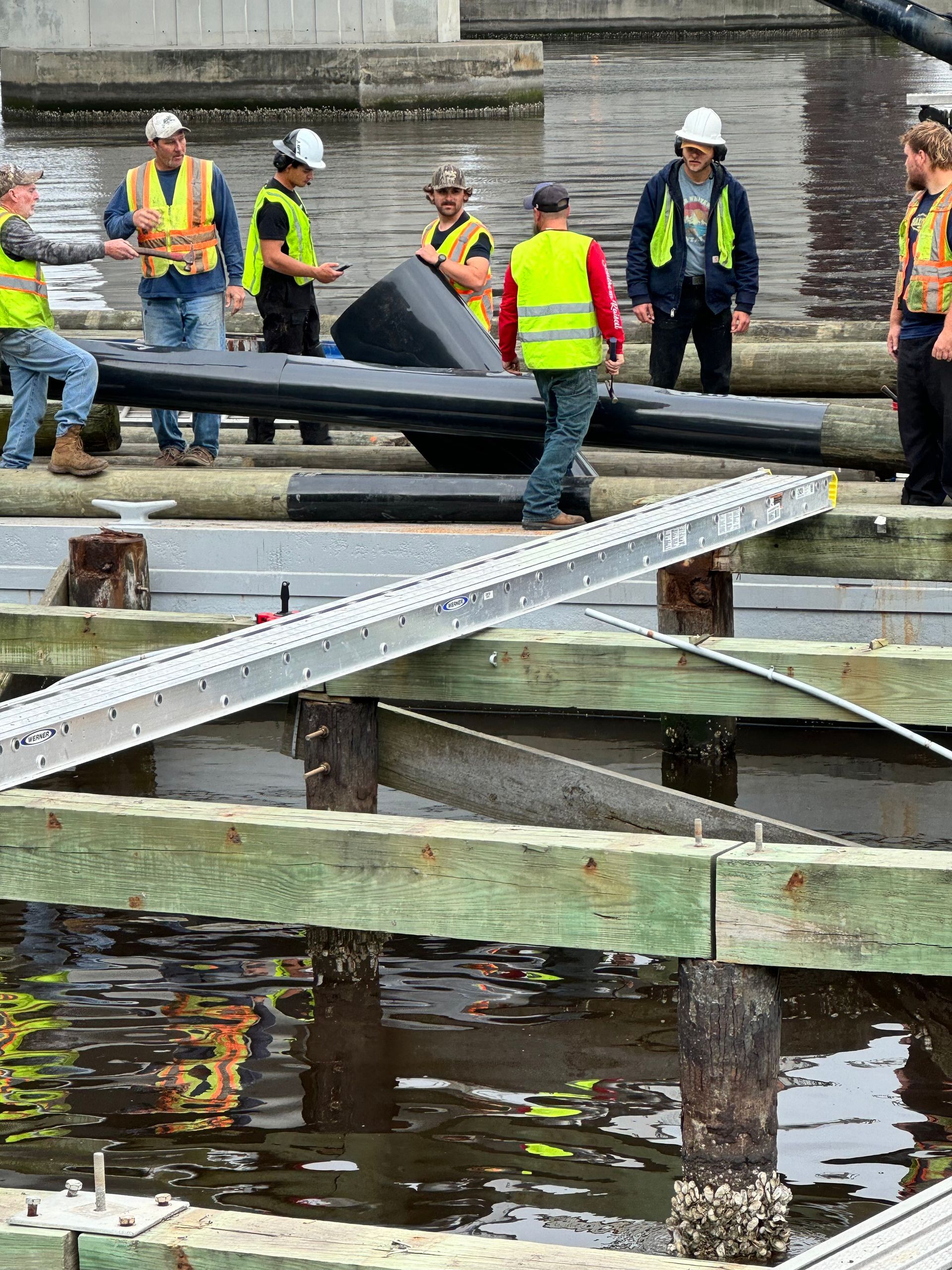 Construction workers on a dock with lumber and equipment. Several wear safety vests and are interacting. Water surrounds pilings.
