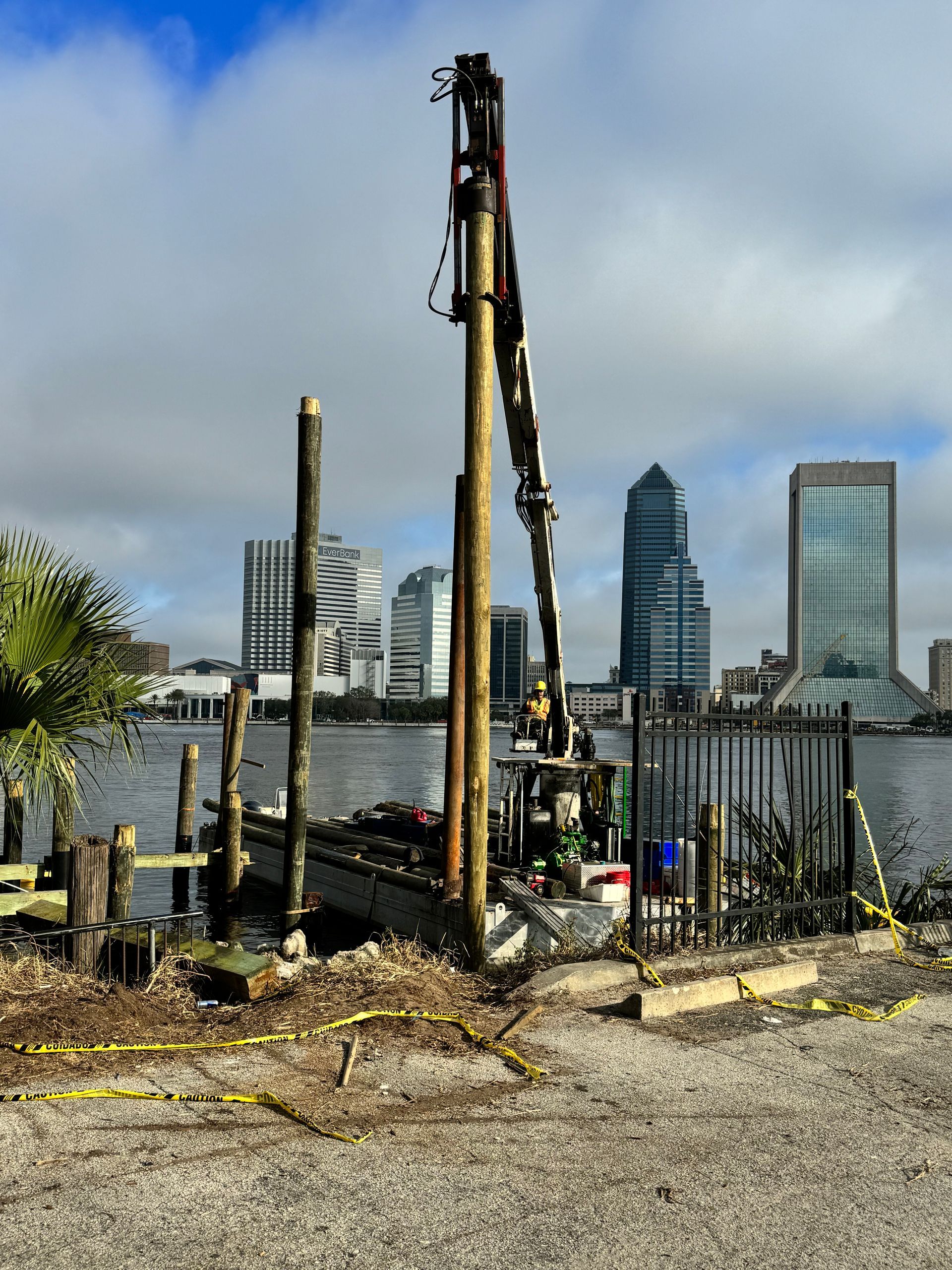 Construction on a pier with a tall machine, cityscape in the background, cloudy sky.