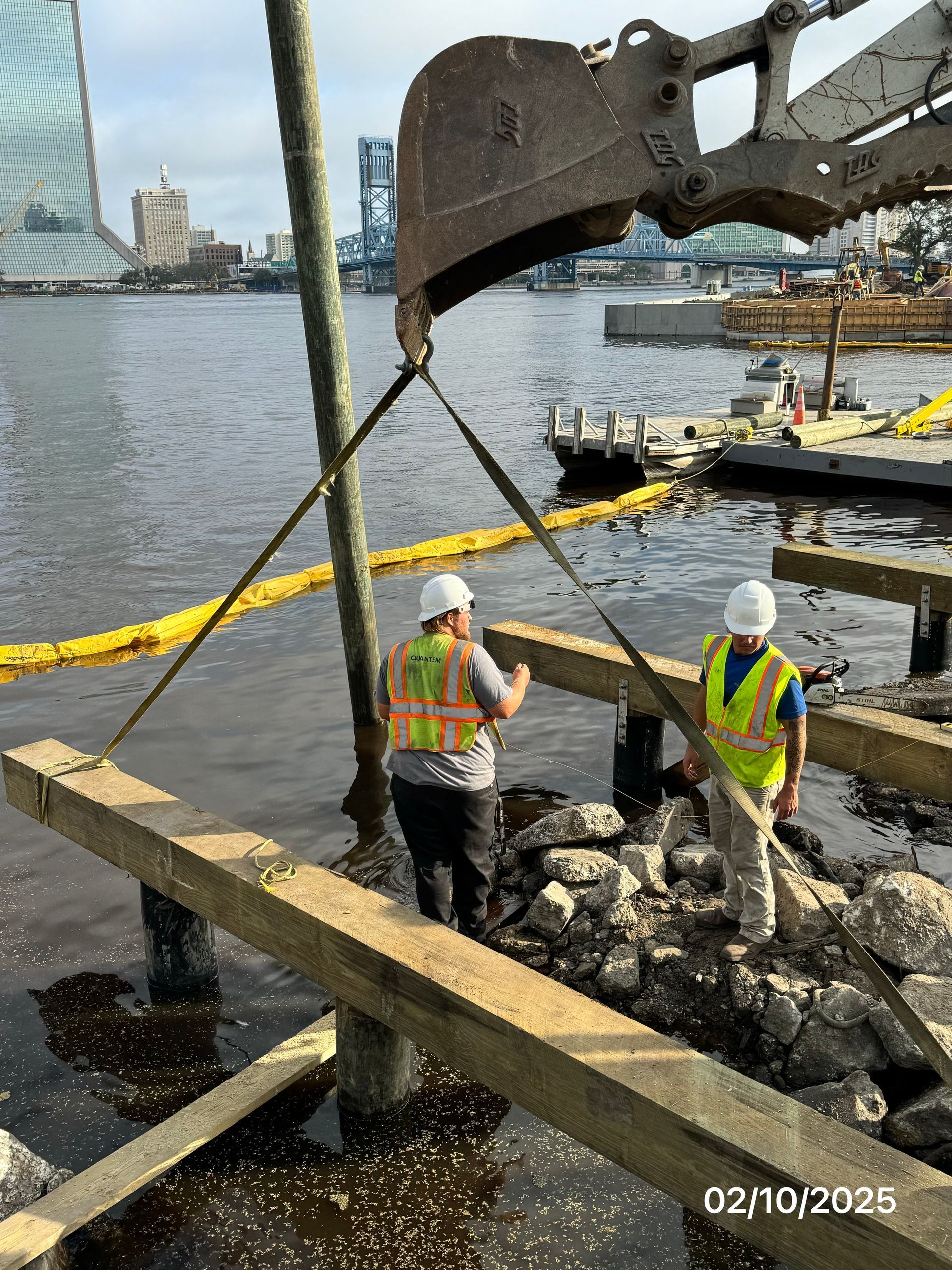 Construction workers installing wooden beams at a waterfront site, excavator overhead.