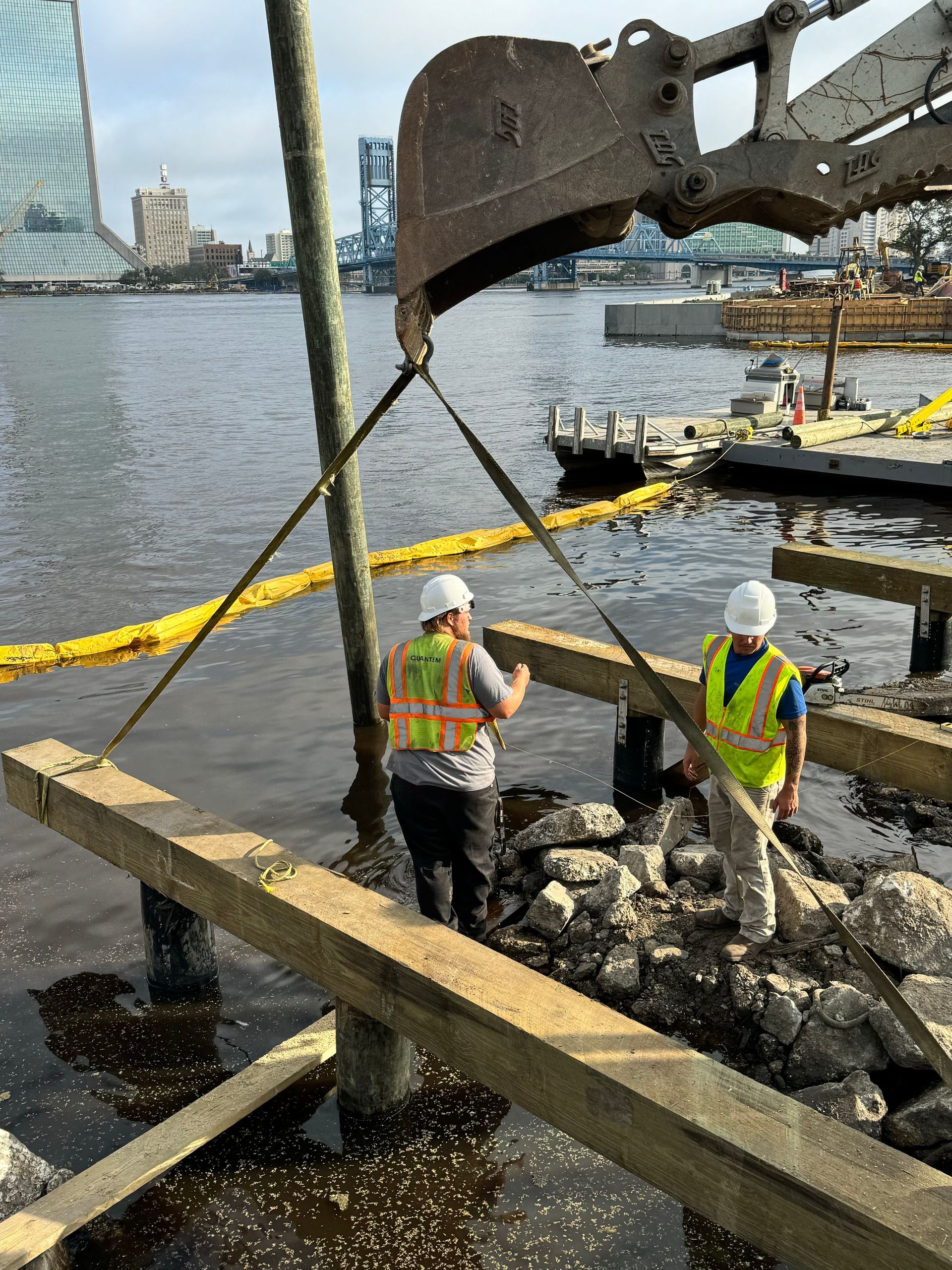 Construction workers near water, with excavator placing a wooden beam.  One worker is inspecting the beam, the other observing.