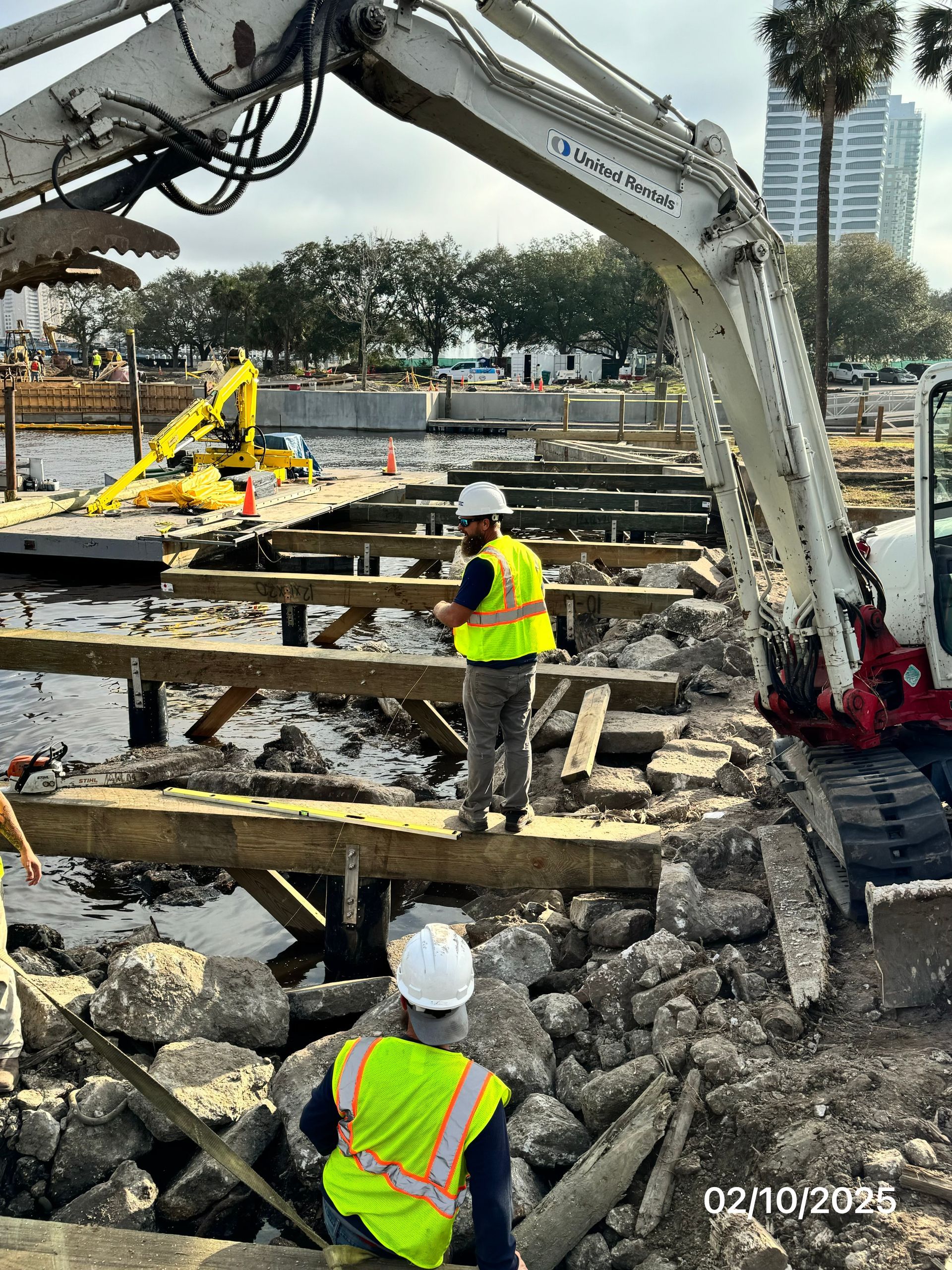 Construction workers removing a wooden dock with an excavator.  One worker stands on the dock. Setting: waterfront.