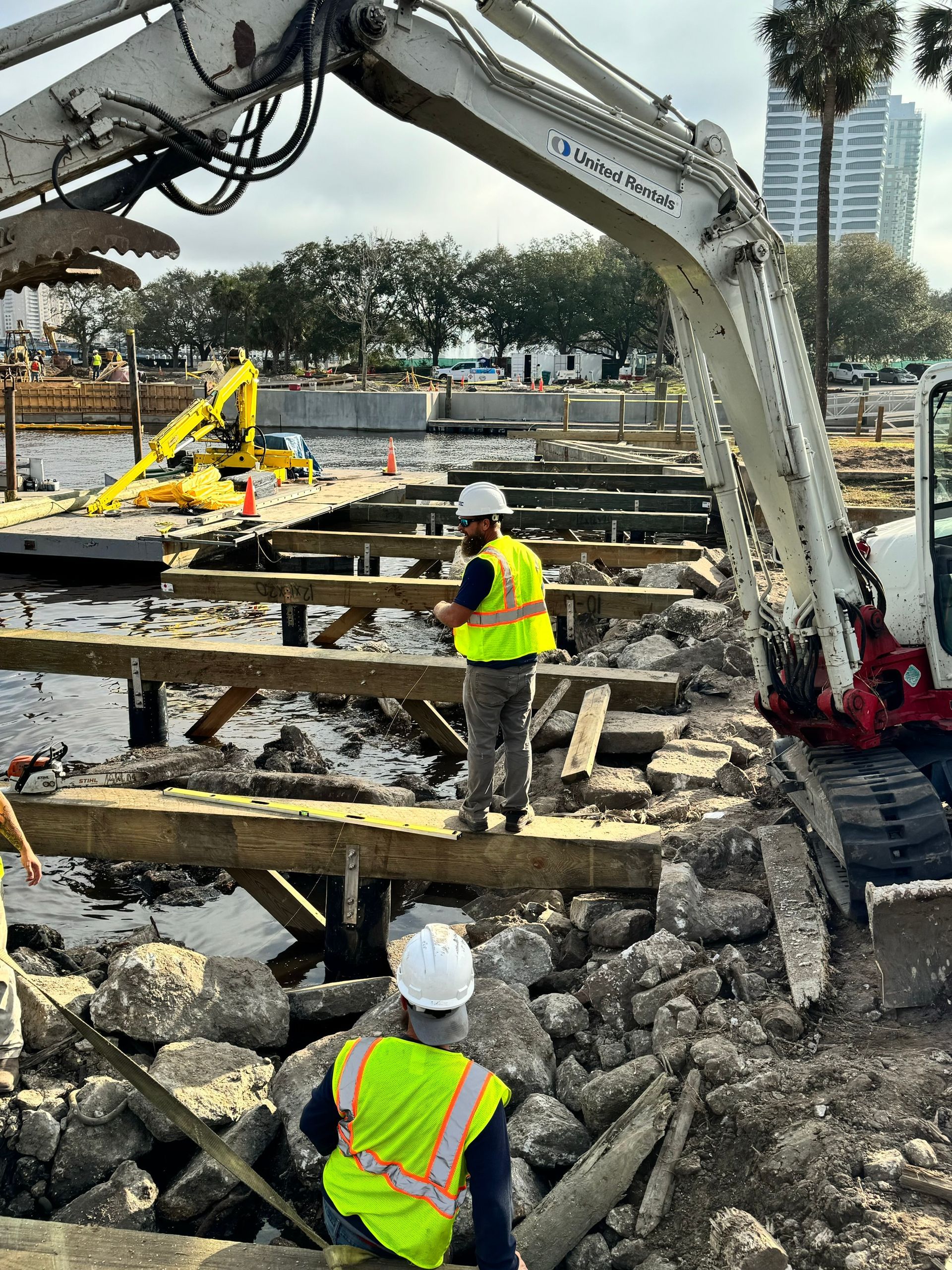 Construction workers dismantling a dock with an excavator near a waterfront.