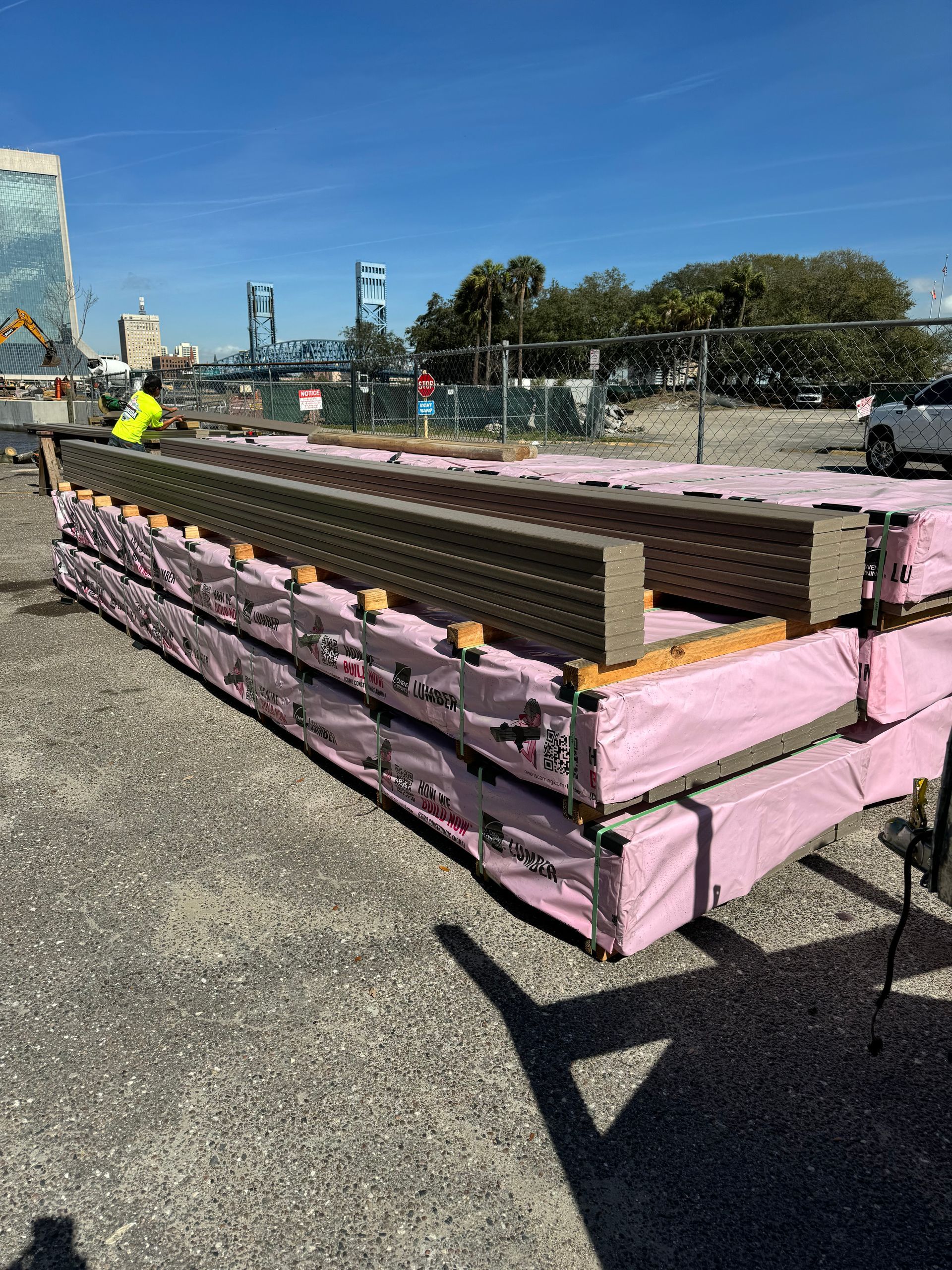 Piles of brown construction materials wrapped in pink plastic. A worker in yellow vest. Construction site. Sunny.