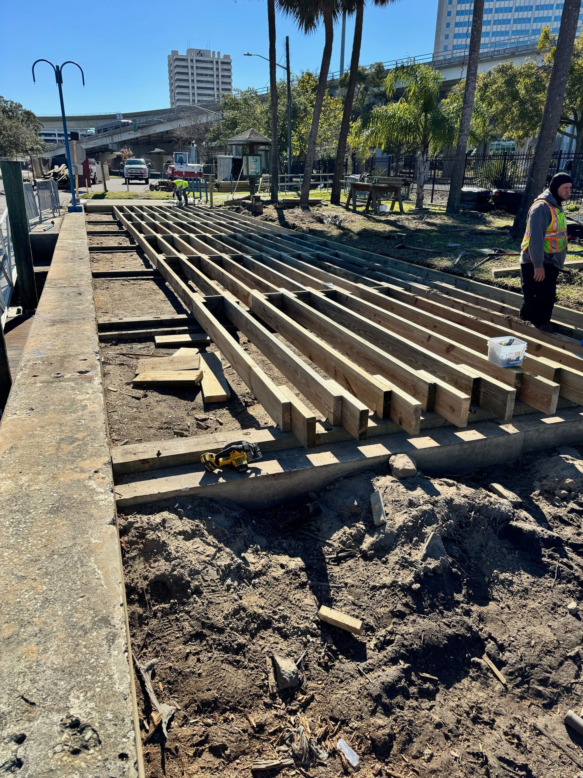 Construction site: wooden beams laid out on dirt ground next to concrete wall. Worker standing. City in background.