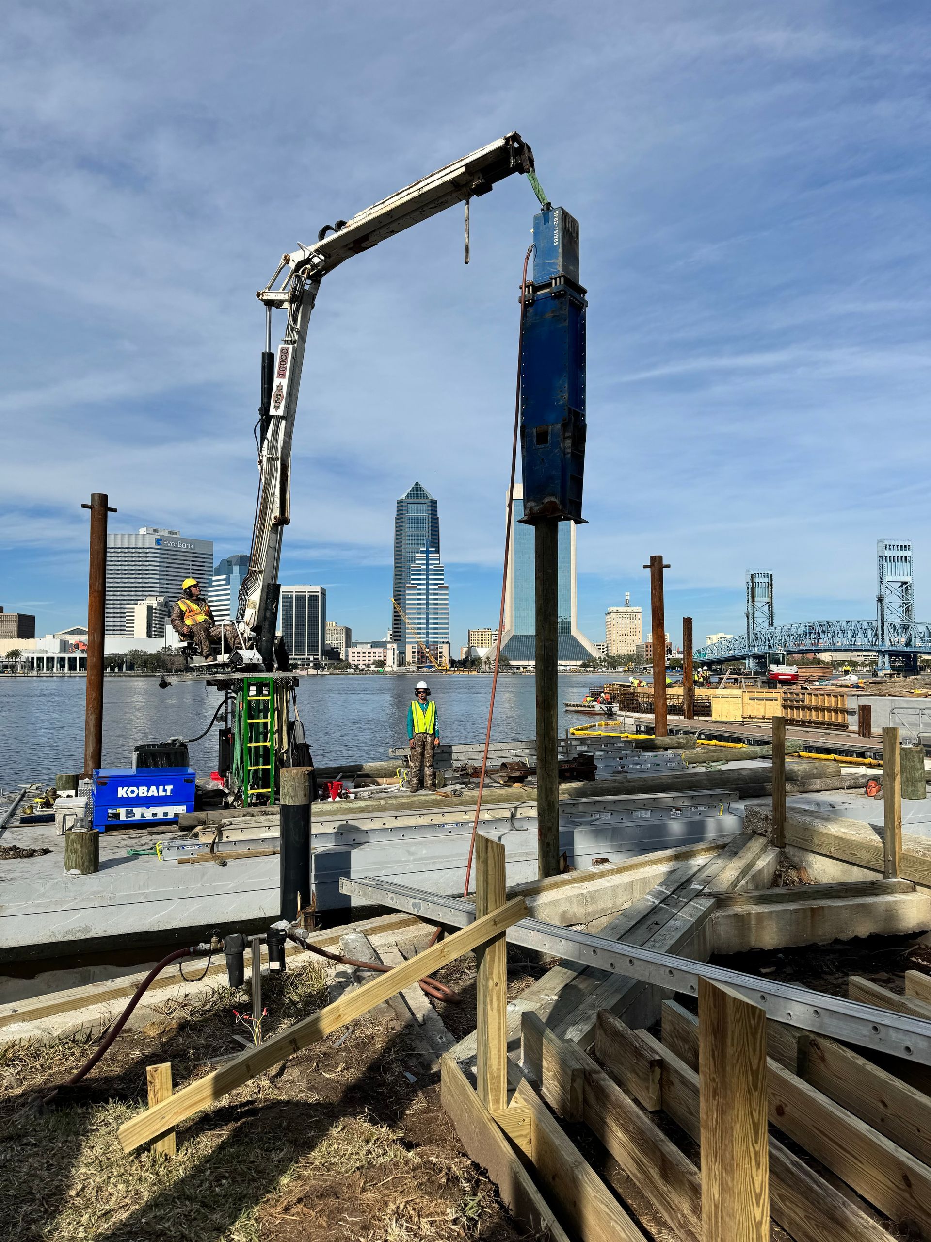 Construction site with a machine driving a pile into the ground, city skyline in background.