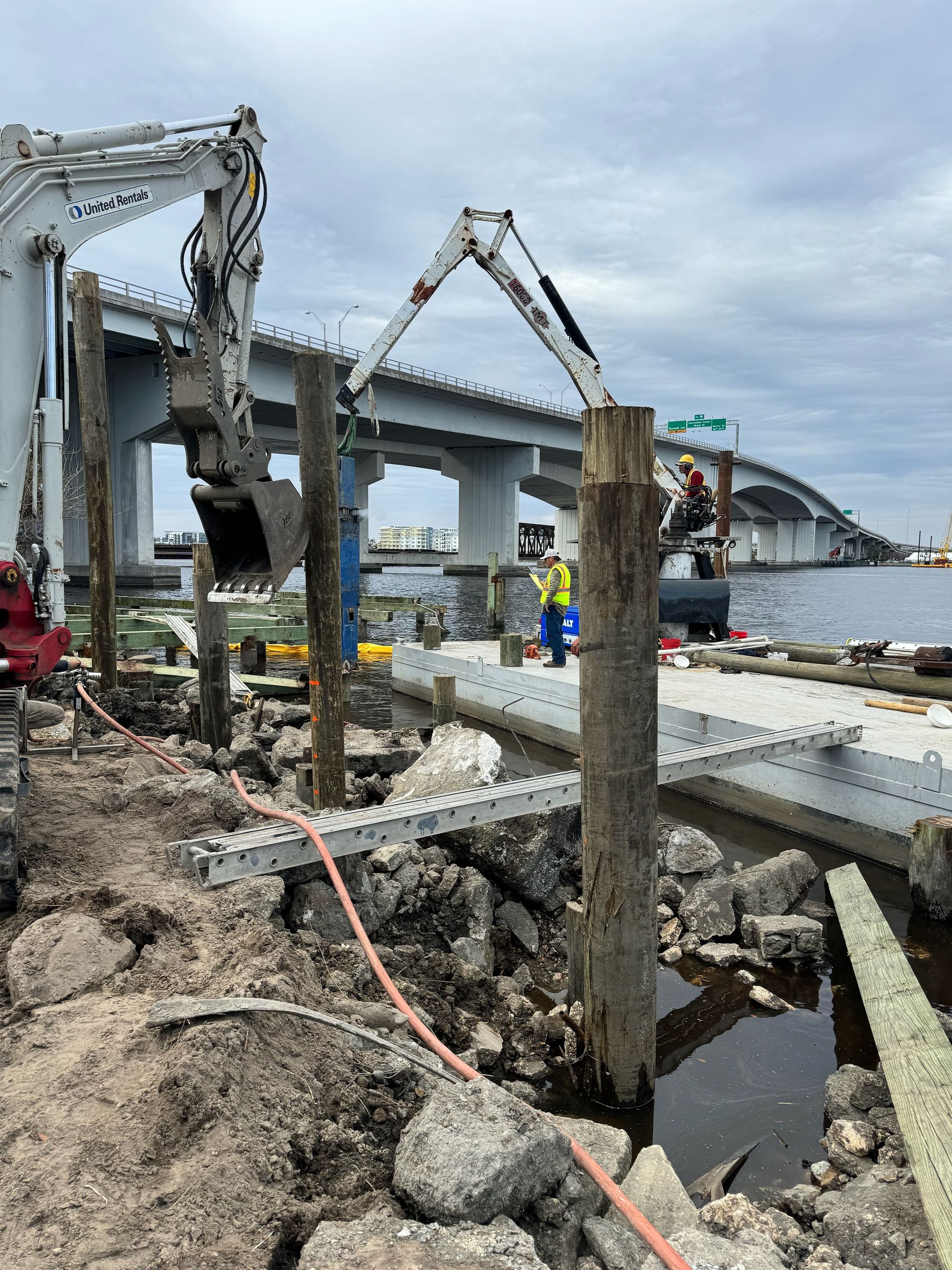 Construction site with excavators removing wooden pilings near water, under a bridge. A worker stands nearby.