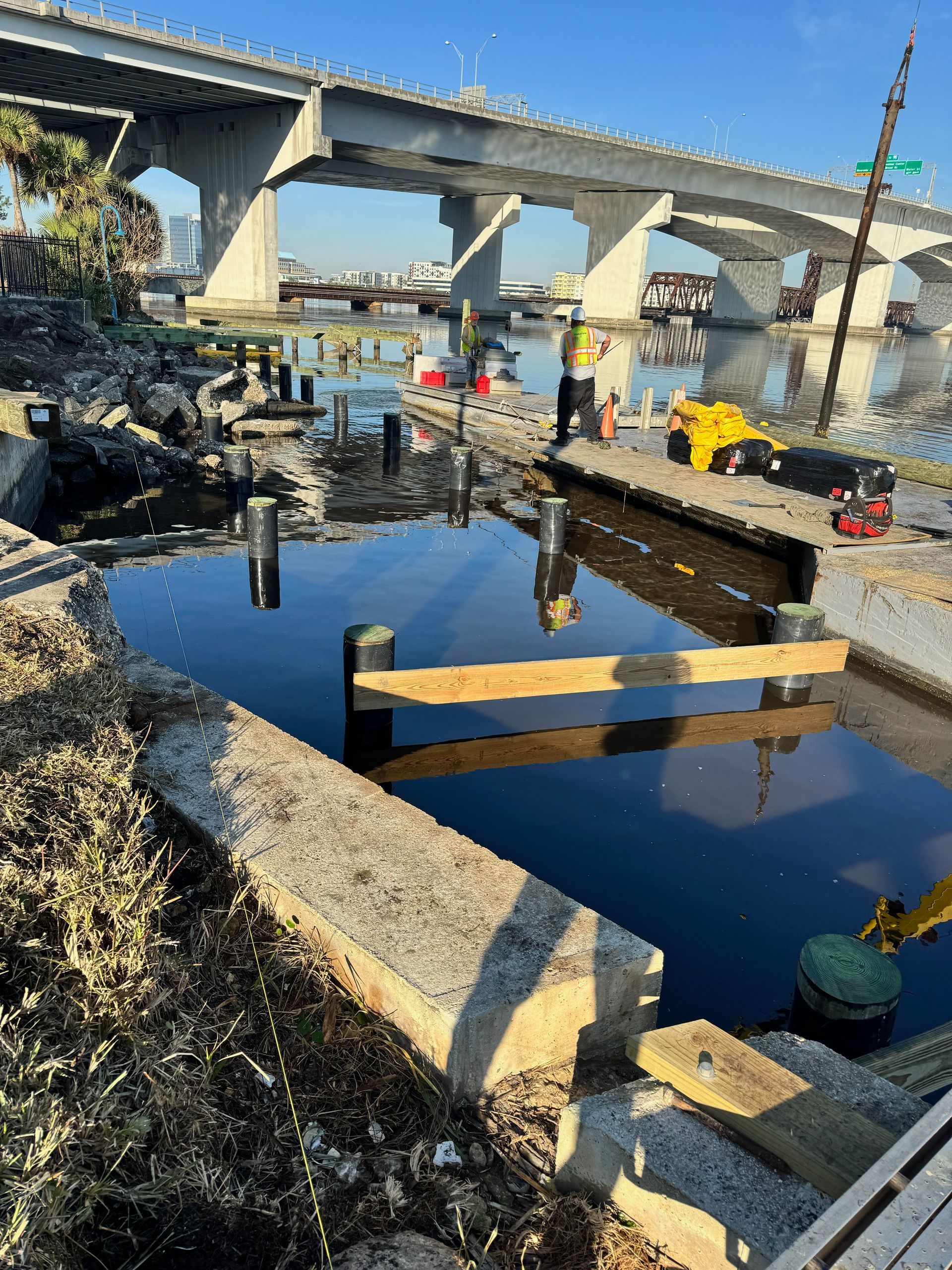 Construction near water with bridge overhead.  Black water and wooden supports are visible.