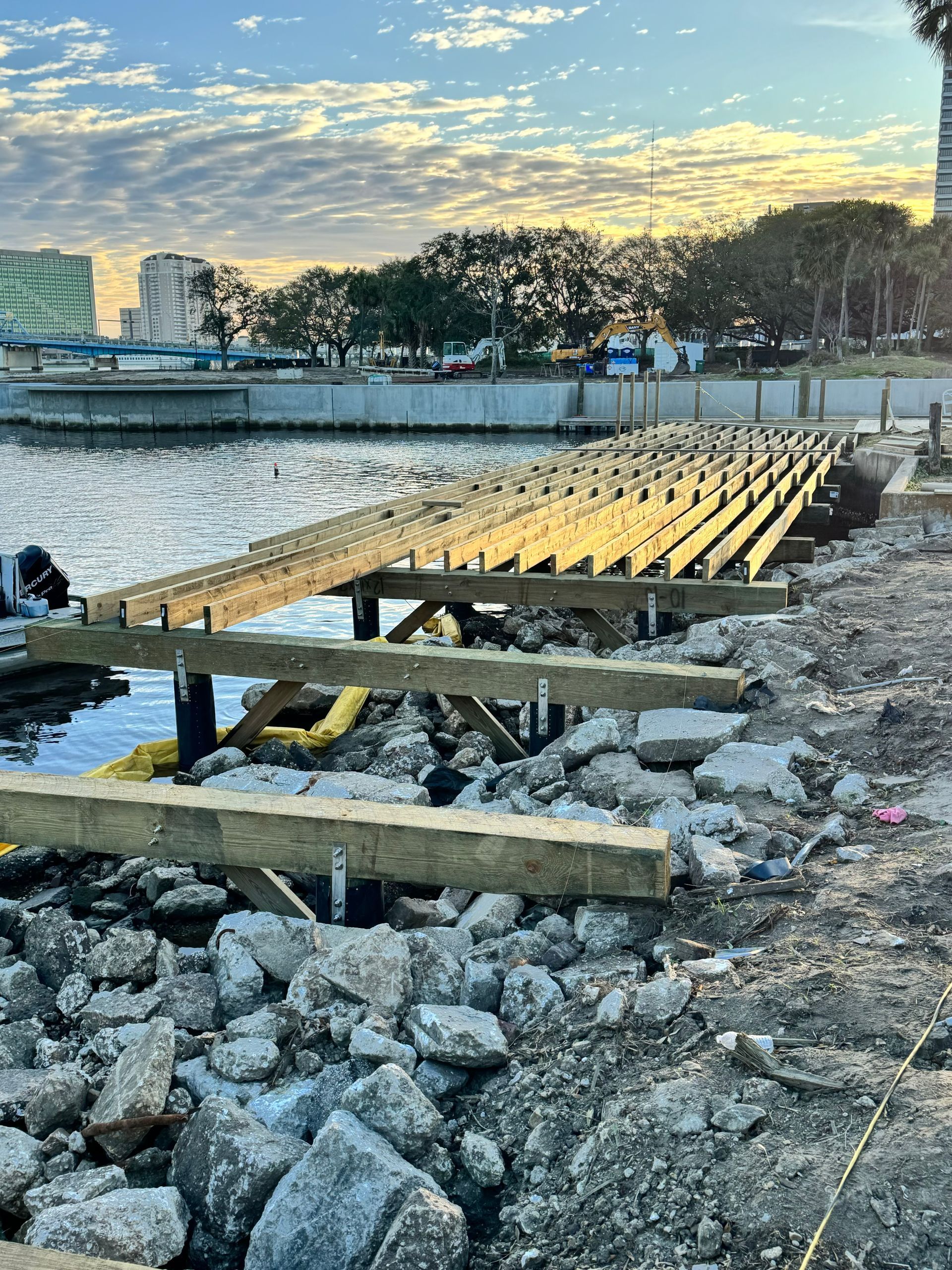 Construction of a wooden dock over water with exposed support beams and rubble in foreground; cityscape in background.