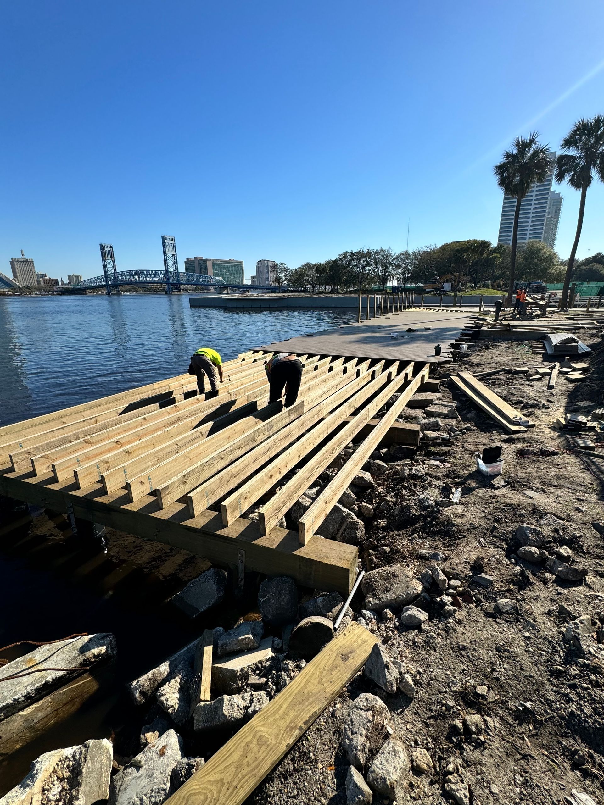 Construction workers rebuilding a pier next to a body of water with a bridge and buildings in the background.