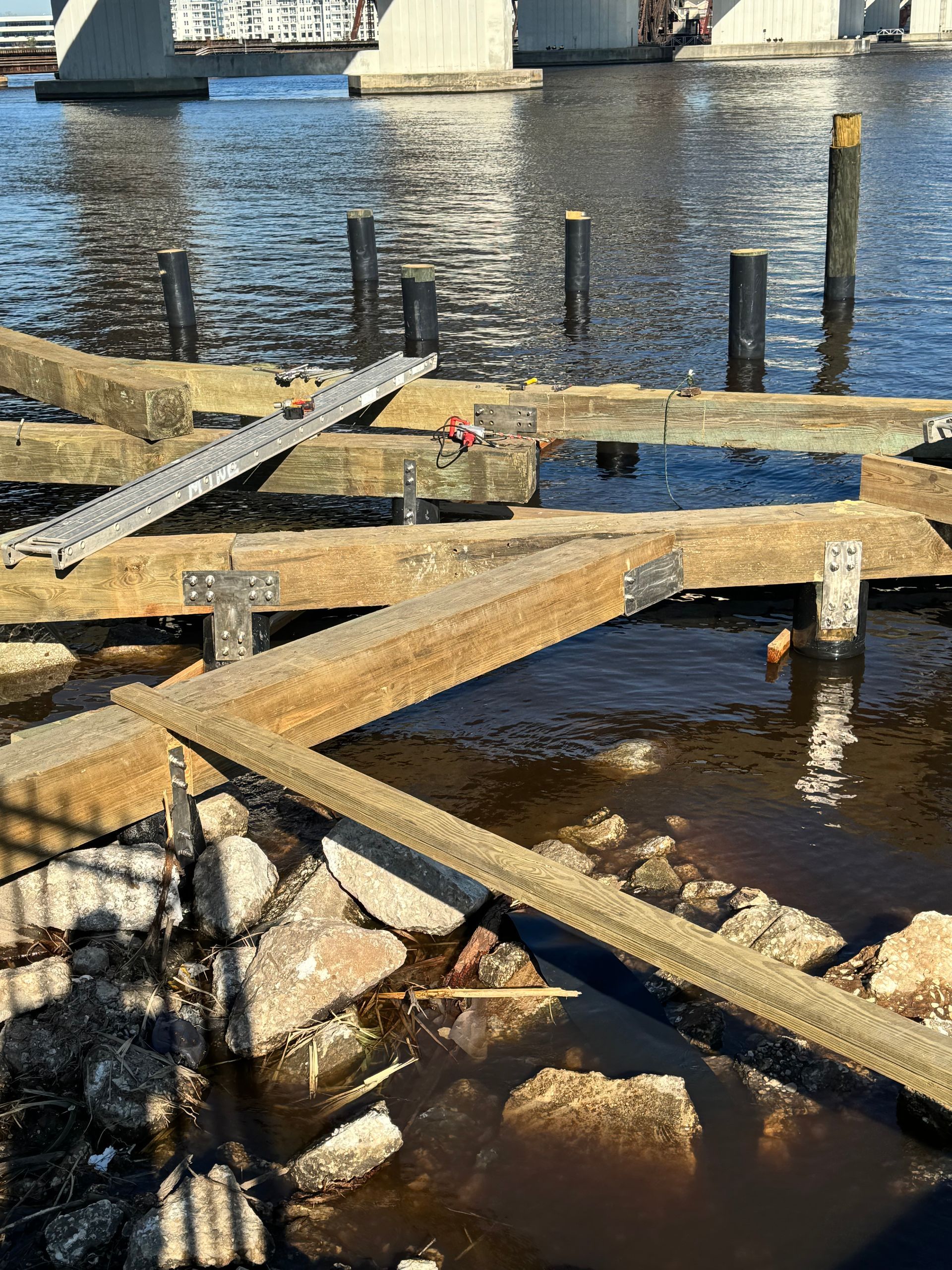 Damaged wooden pier in water, with metal brackets and pillars under a bridge.