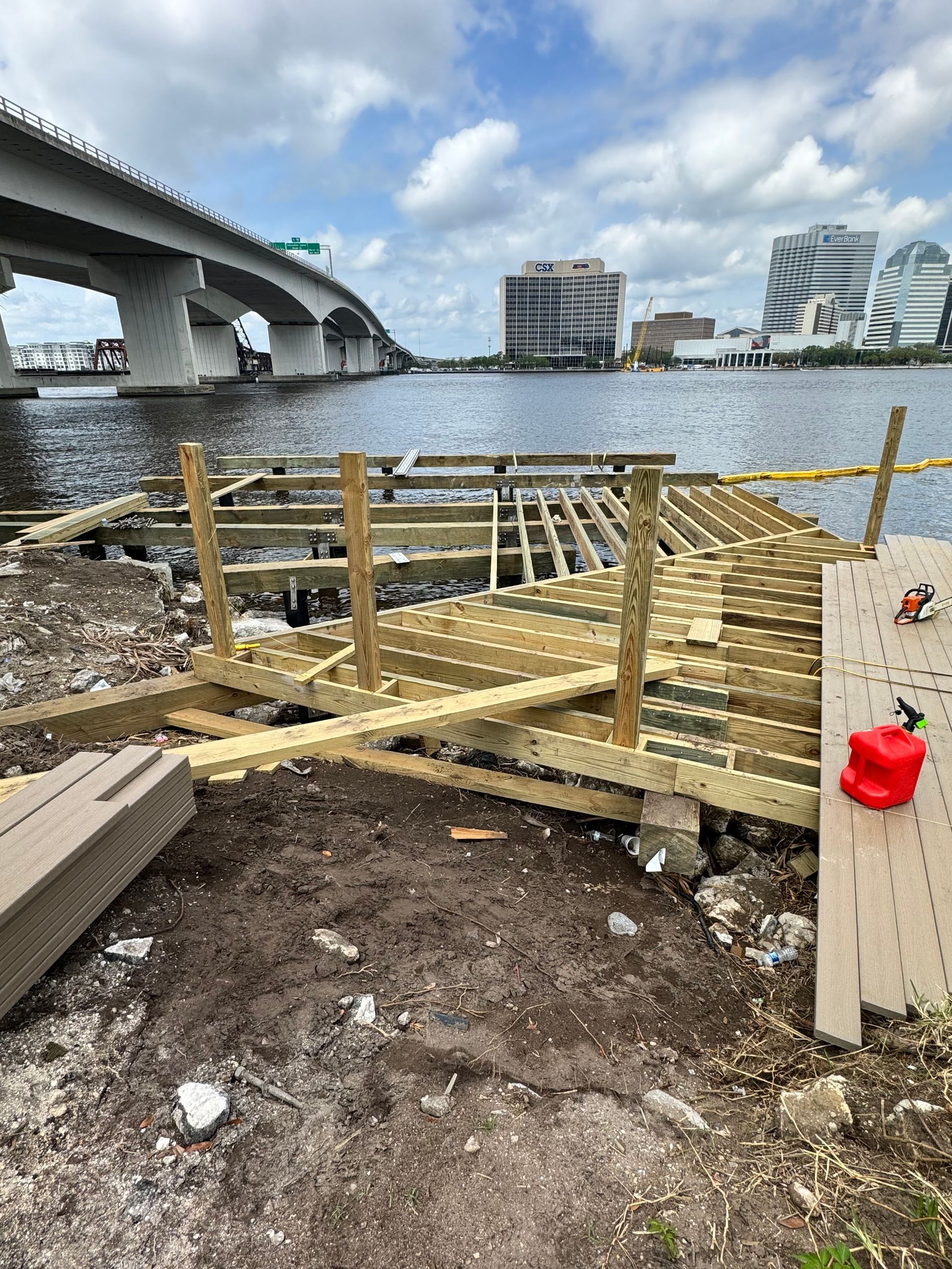 Wooden dock under construction on a river with a bridge and city skyline in the background.