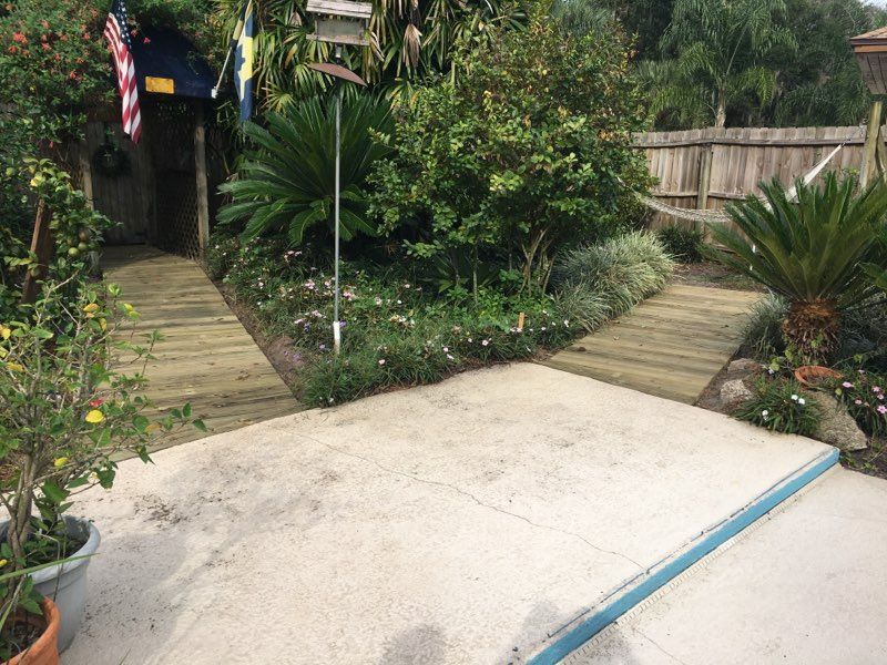 Wooden paths diverge from a concrete patio into a garden with lush greenery and a small wooden structure.