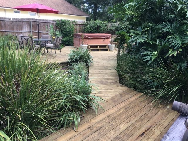 Wooden deck pathway leads to a hot tub. Outdoor dining set and lush plants are visible.