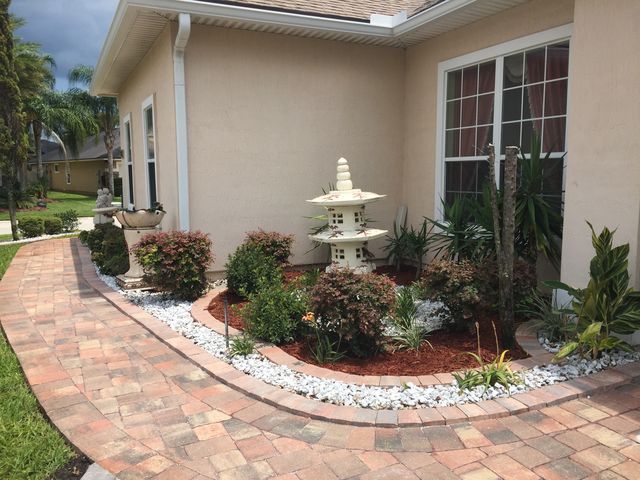 Brick pathway curves past a tan house and garden beds with white rocks, bushes, and a pagoda statue.