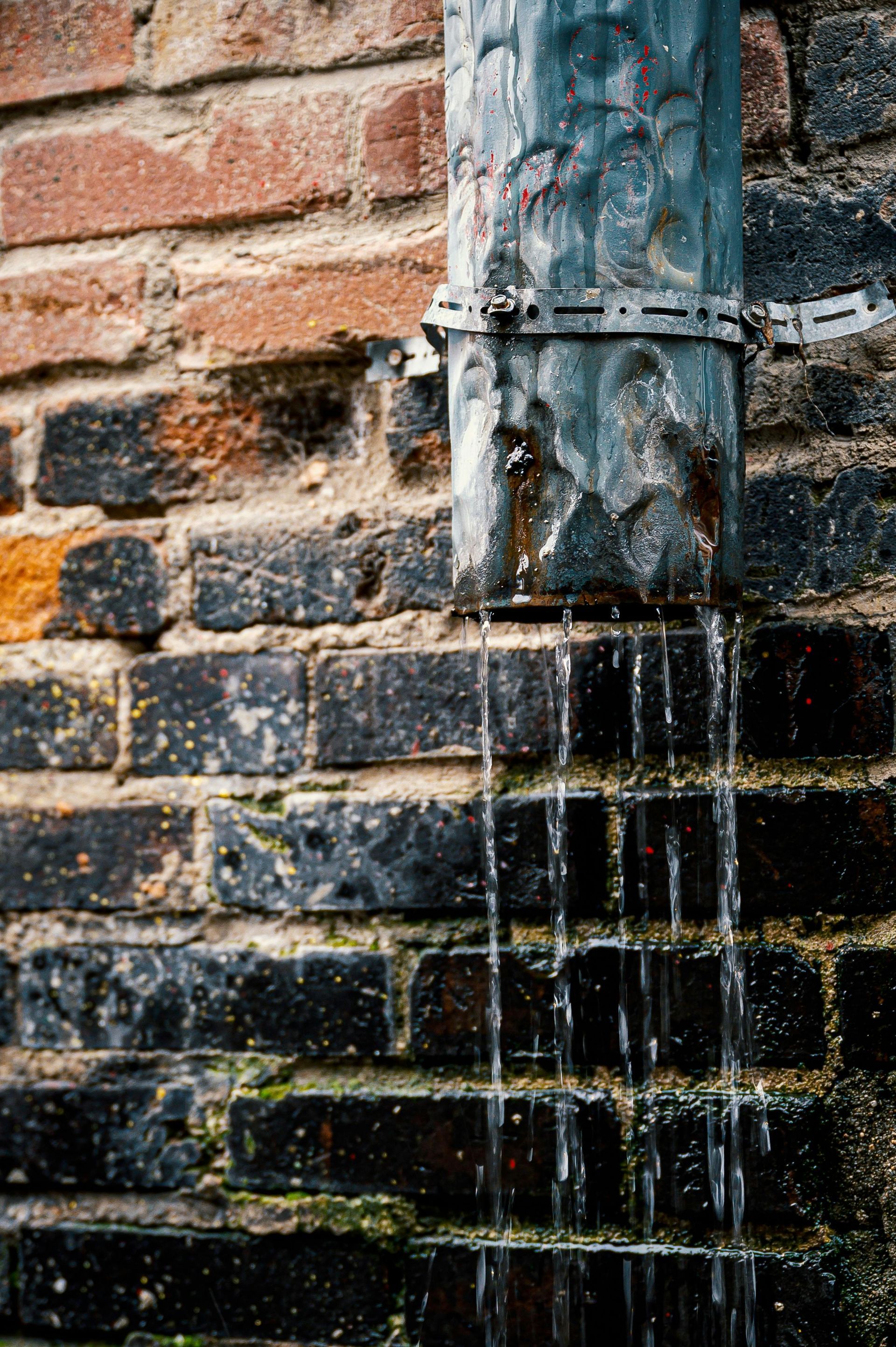 Rainwater pouring from a weathered metal downspout against a dark, brick wall.