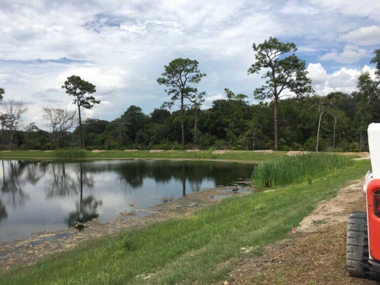 Pond surrounded by green grass, trees, and cloudy sky. A small white and orange vehicle is on the right.