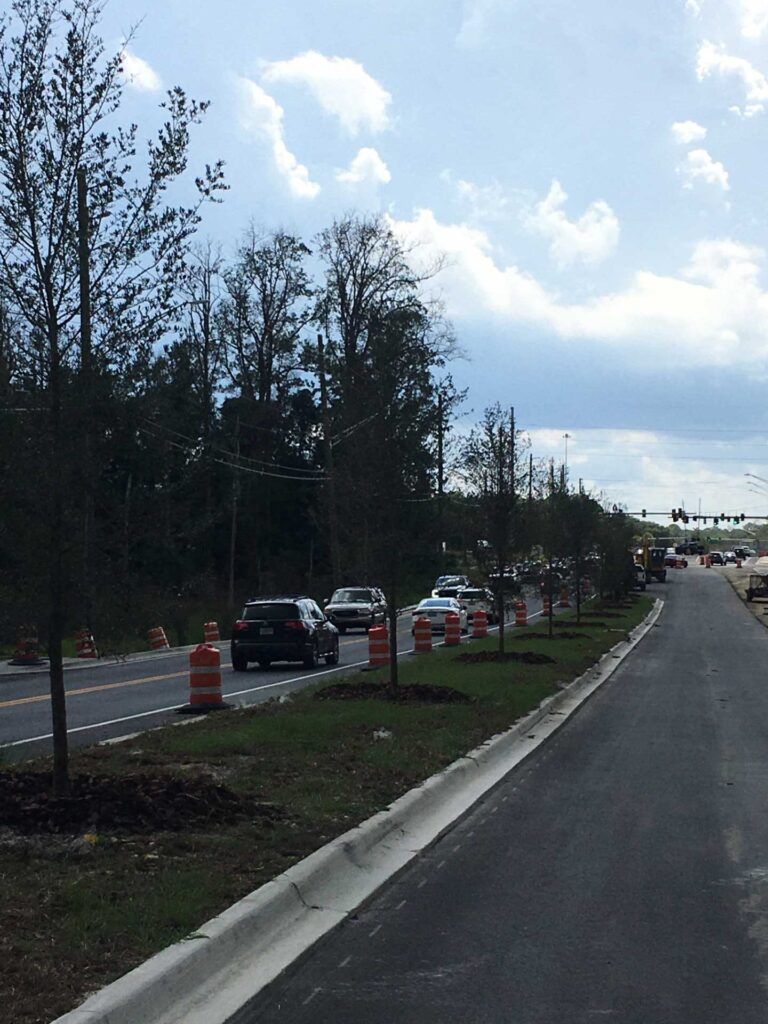 Road under construction, cars driving, orange cones, trees along the side. Cloudy sky in the background.