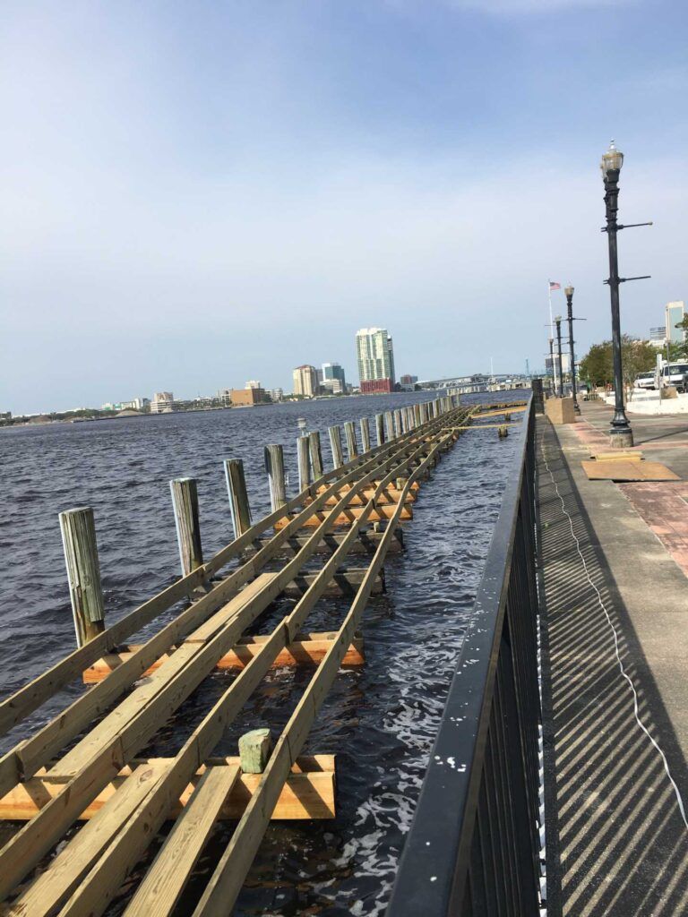 Wooden pier under construction along a waterfront, with city skyline in the distance and a sidewalk on the right.