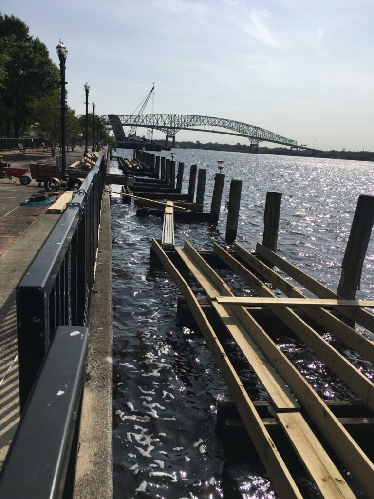 Waterfront pier under construction with a bridge in the background. Wooden beams in the water along a walkway.