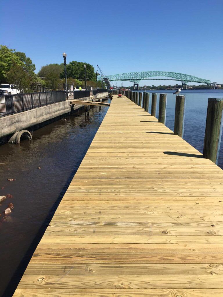 A wooden pier extends into water, with a green bridge in the background. Sunny day.