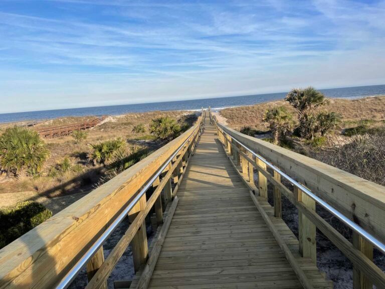 Wooden boardwalk to a beach with ocean view, blue sky, and surrounding dunes.