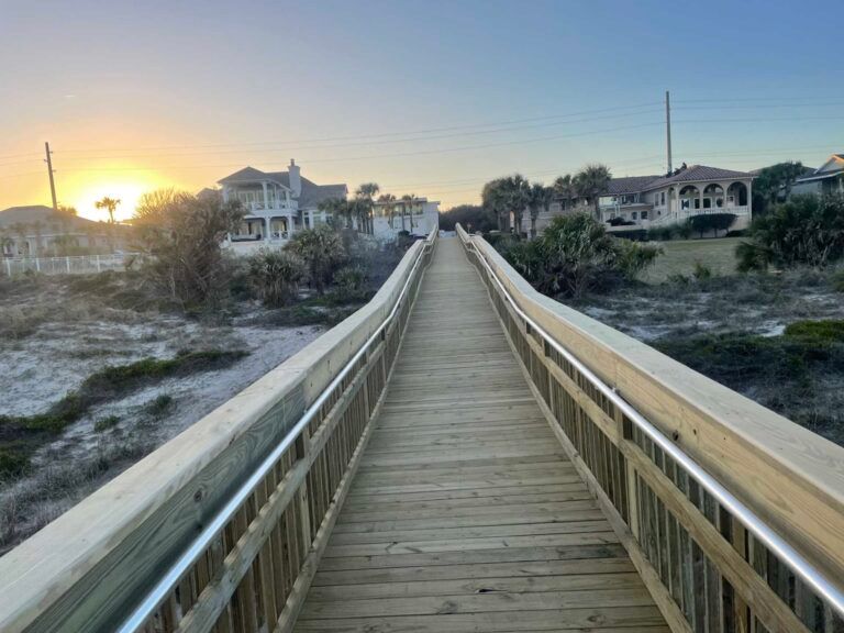 Wooden boardwalk leading over dunes towards beach houses at sunset.