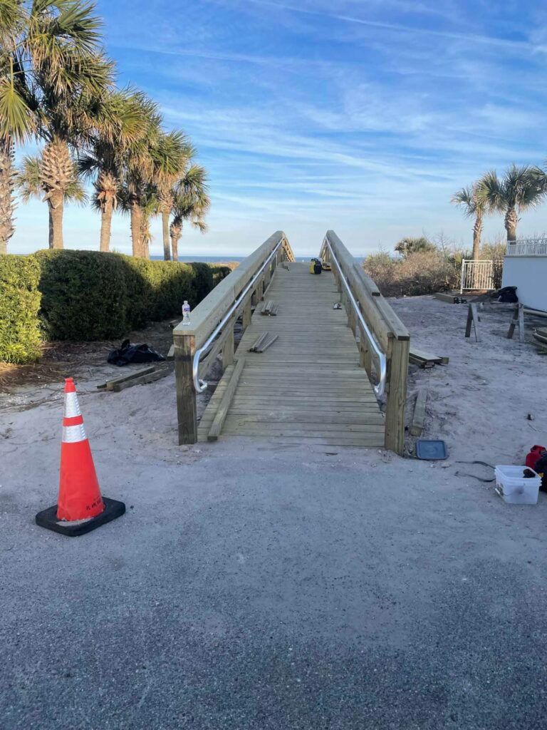Wooden boardwalk leading to a beach, with palm trees and a blue sky. A traffic cone is in the foreground.