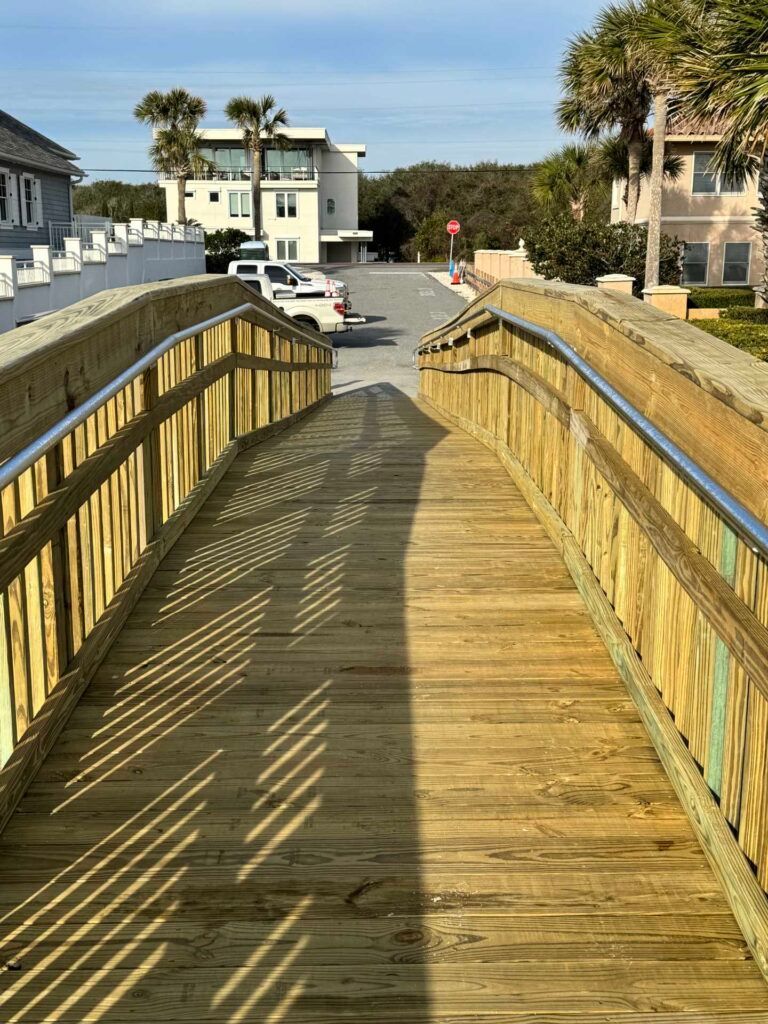 Wooden bridge over a road; buildings and palm trees in background; sunny day.