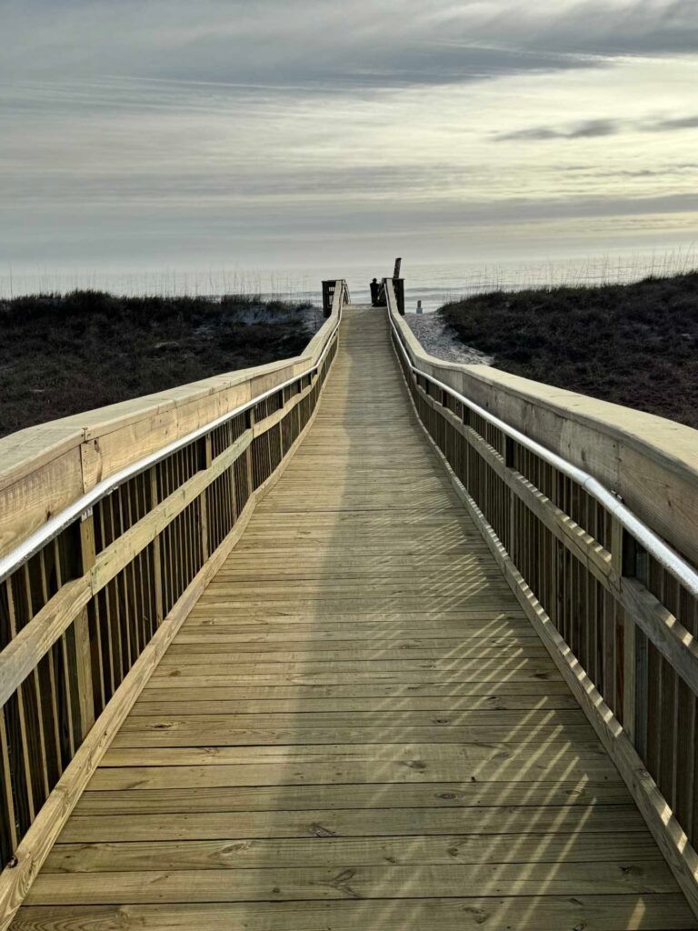 Wooden boardwalk leading to the beach, with ocean and cloudy sky in the background.