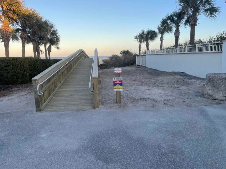 Wooden ramp leading to beach, with signs posted. Palm trees and low wall in the background.
