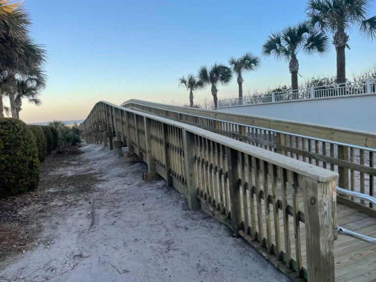 Wooden boardwalk with railing leads over sand towards beach and palm trees.