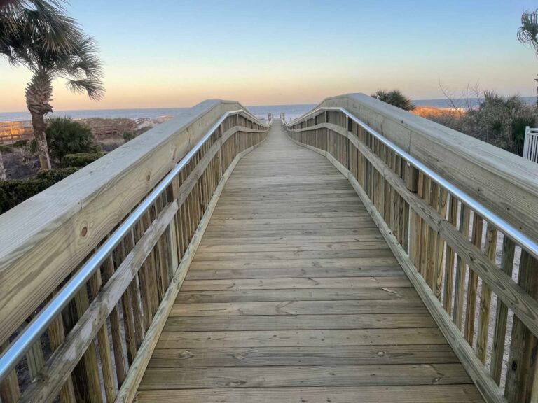 Wooden boardwalk leads to the ocean at sunset, with handrails on either side.