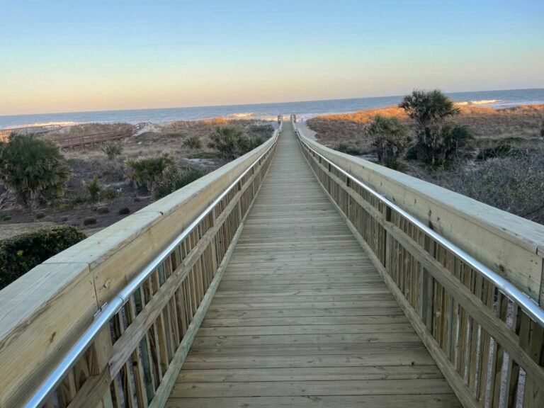 Wooden boardwalk leading to a beach and ocean; bright sky overhead.