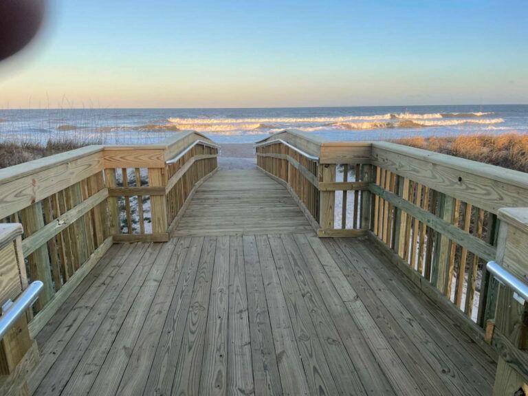 Wooden boardwalk leading to the beach and ocean. Blue sky, waves in the distance.