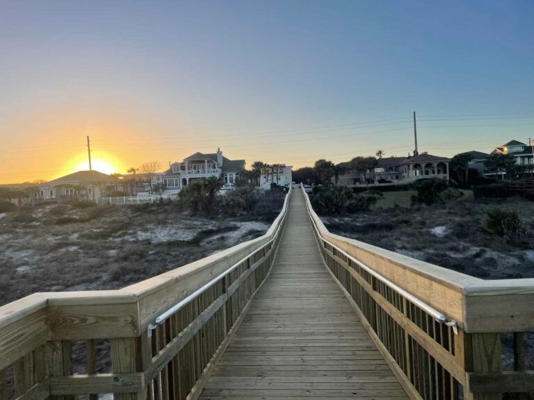 Wooden boardwalk leading towards the horizon at sunset, with buildings visible in the distance.