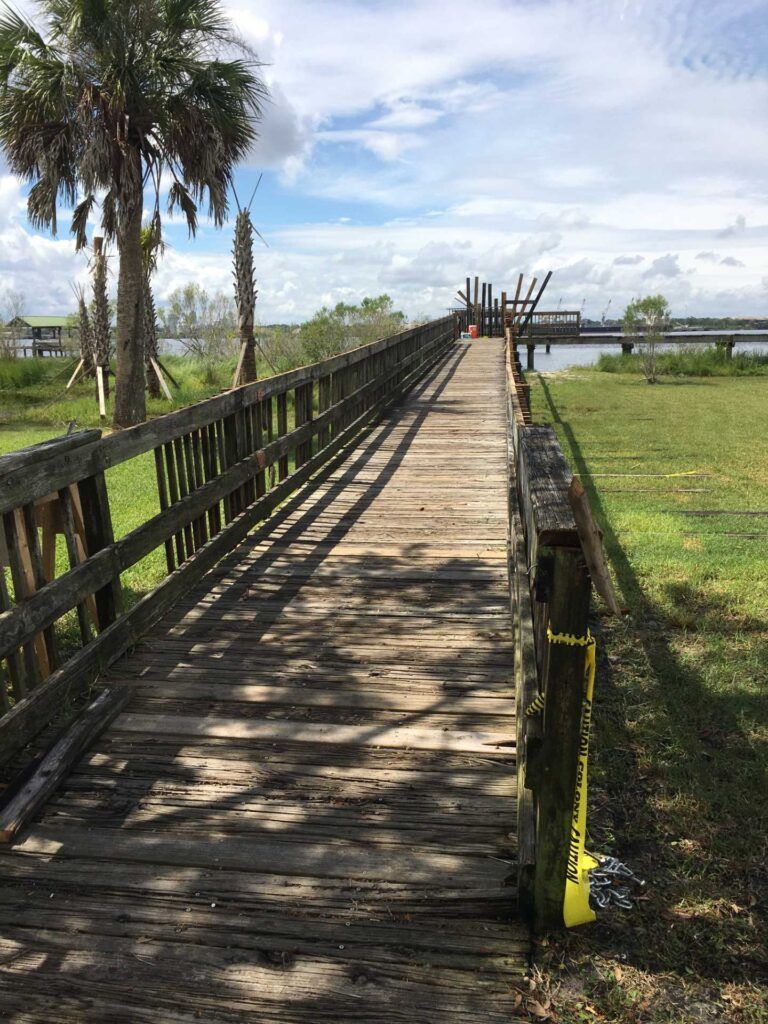 Wooden boardwalk extends toward water, with palm trees and a grassy area under a sunny sky.