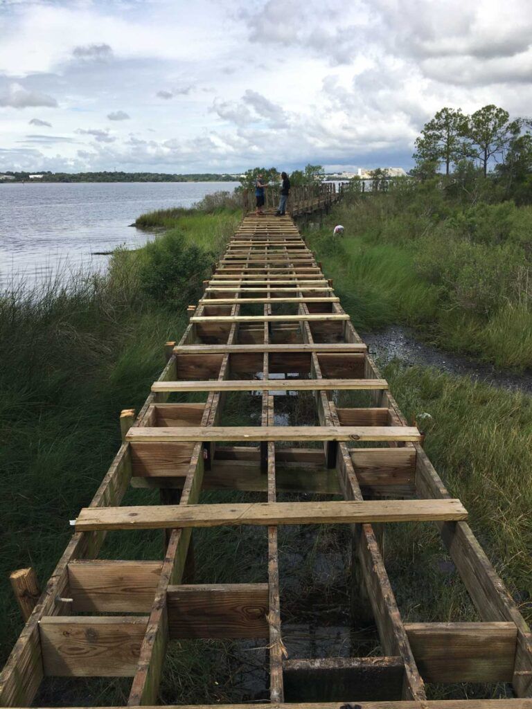 Wooden walkway under construction next to a body of water with two figures standing in the distance.