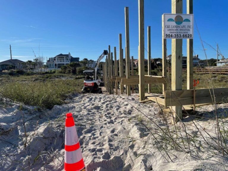 Construction on sandy beach; wooden posts being built with excavator, orange cone in foreground.