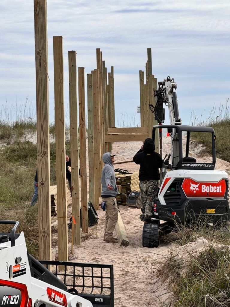 Construction workers building a wooden walkway on a sandy beach. A Bobcat excavator is present.