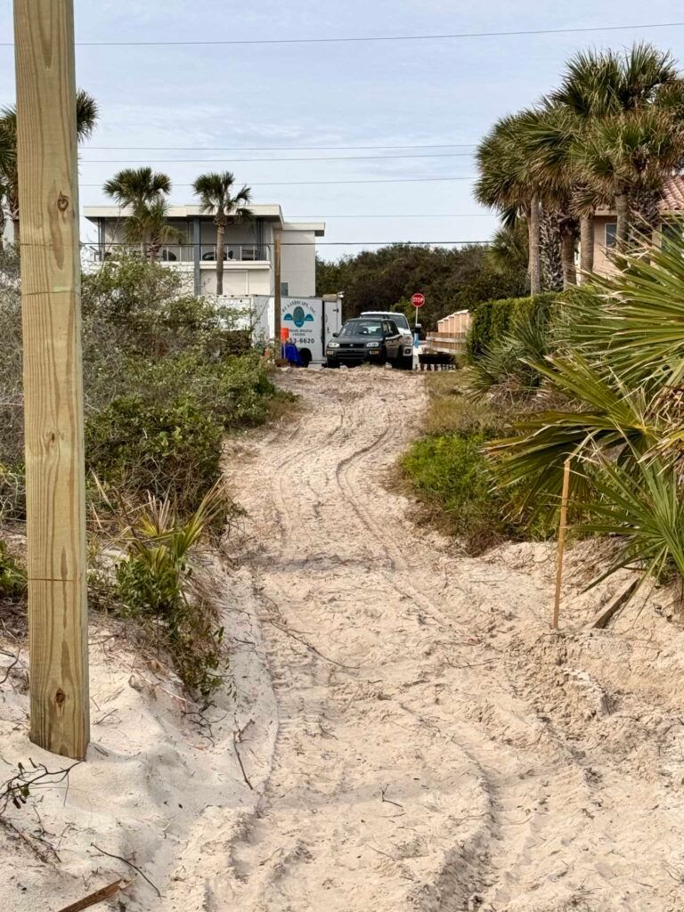 Sandy path leading to a beach house with parked cars. A utility pole is in the foreground.