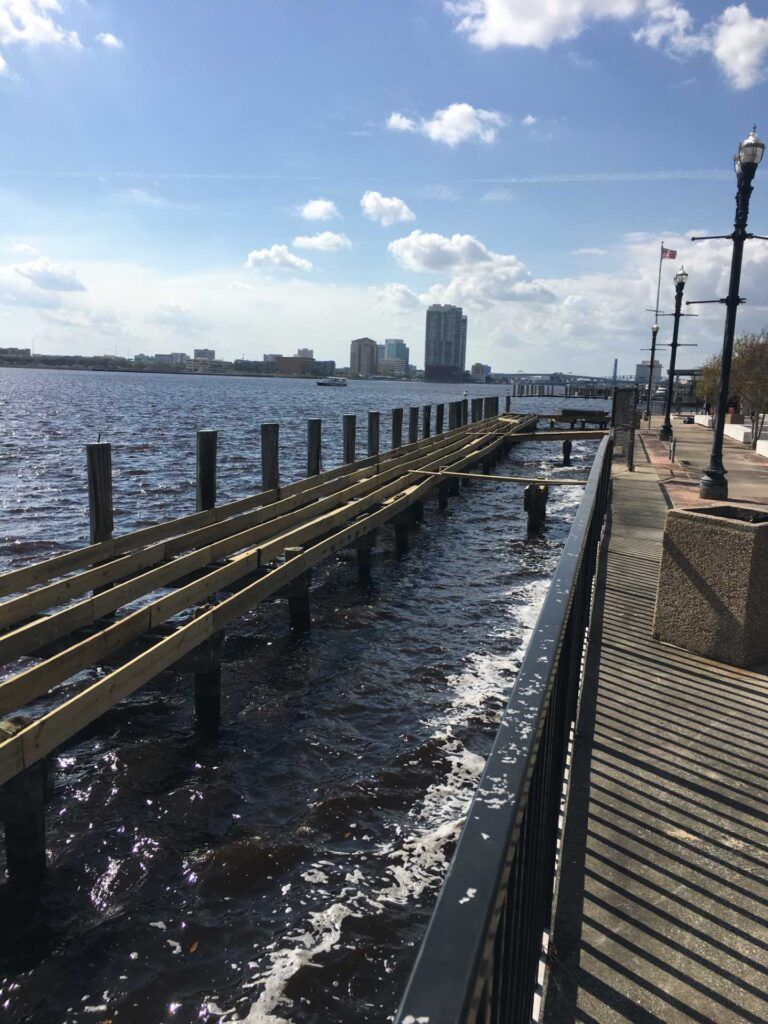 A wooden pier extends into dark water, beside a walkway with lampposts. City skyline visible in the distance.