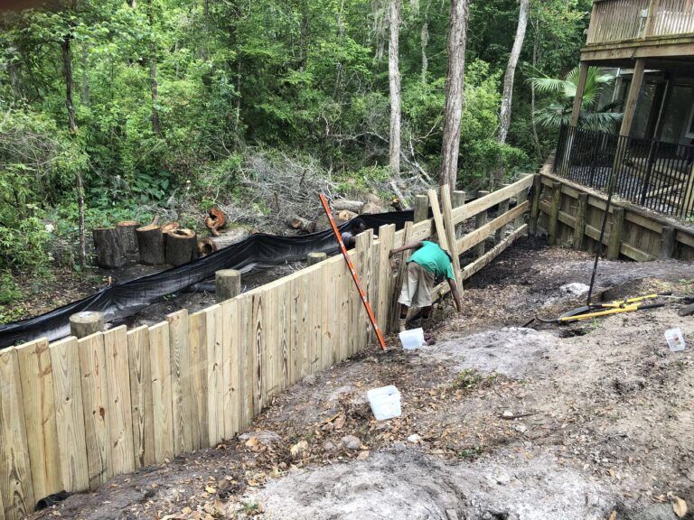 Man building a wooden fence in a wooded area. Black fabric laid down with tree stumps nearby.