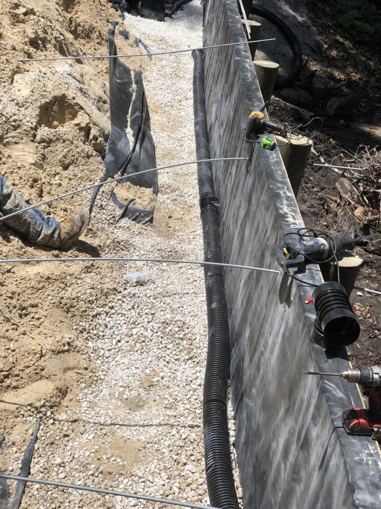 Construction of a retaining wall. Grey concrete, gravel, black drain pipe, and wire rods are visible.