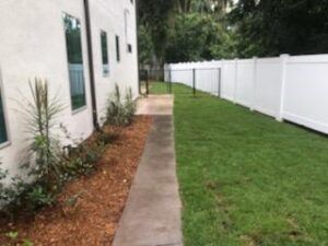 Narrow concrete path beside white house and fence, with mulch border and green lawn in backyard